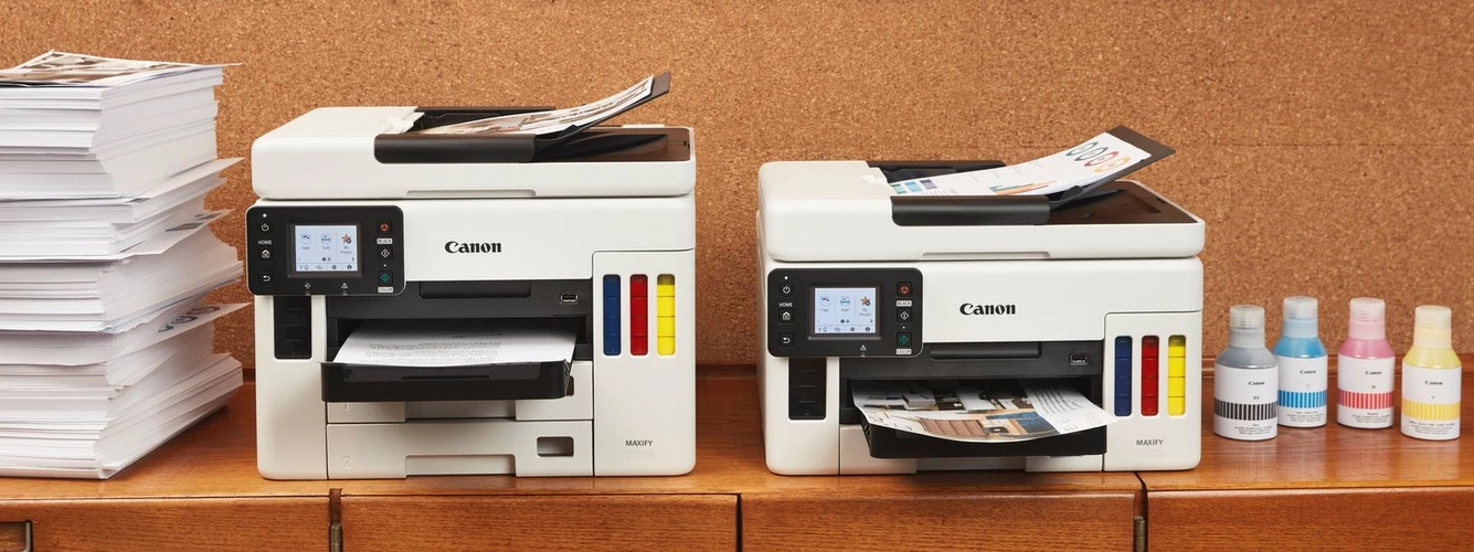 Twee Canon MegaTank-printer op een houtenkast met daarnaast een stapel papier en vier inktflessen